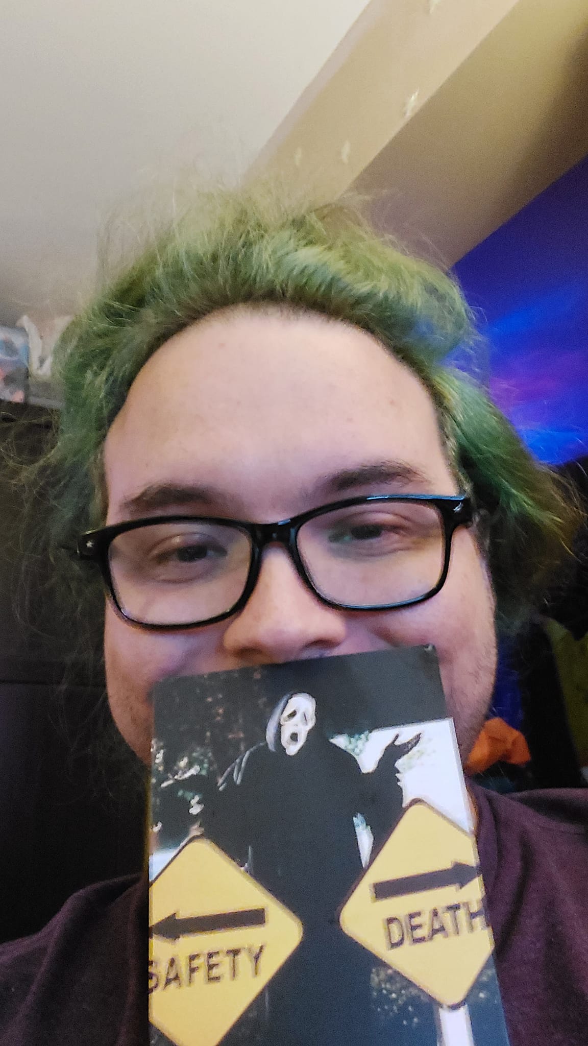 A man with long green hair and no beard holding a post card with a person in a scream mask on it